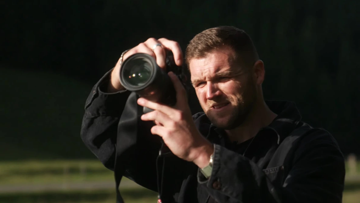 Photographer holding binoculars to his face against a dark background.