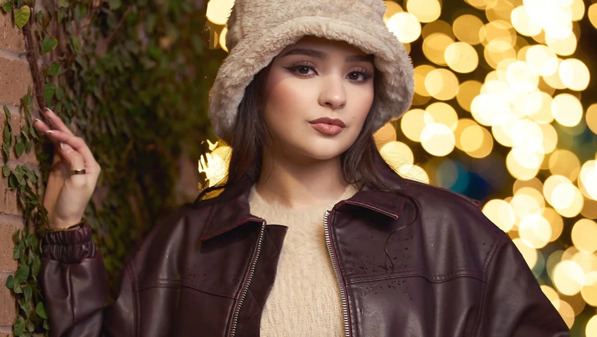 Young woman in brown leather jacket and beanie leaning against ivy-covered wall with golden bokeh lights behind her.