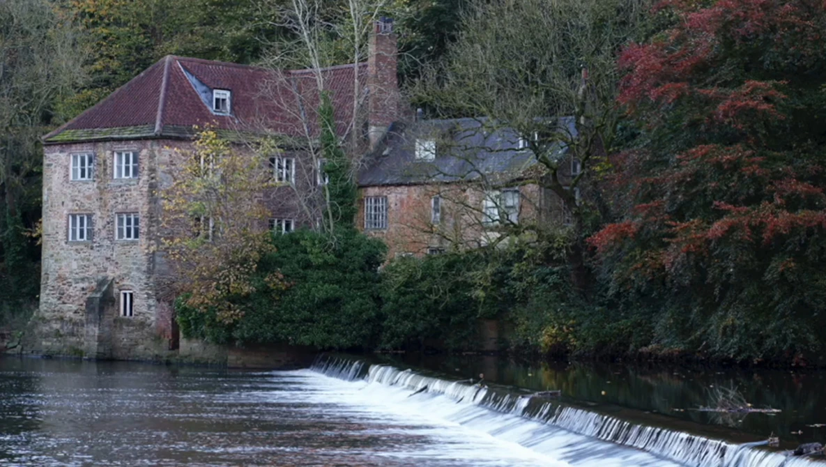 Historic stone mill building beside a river weir with autumn foliage in soft natural light.