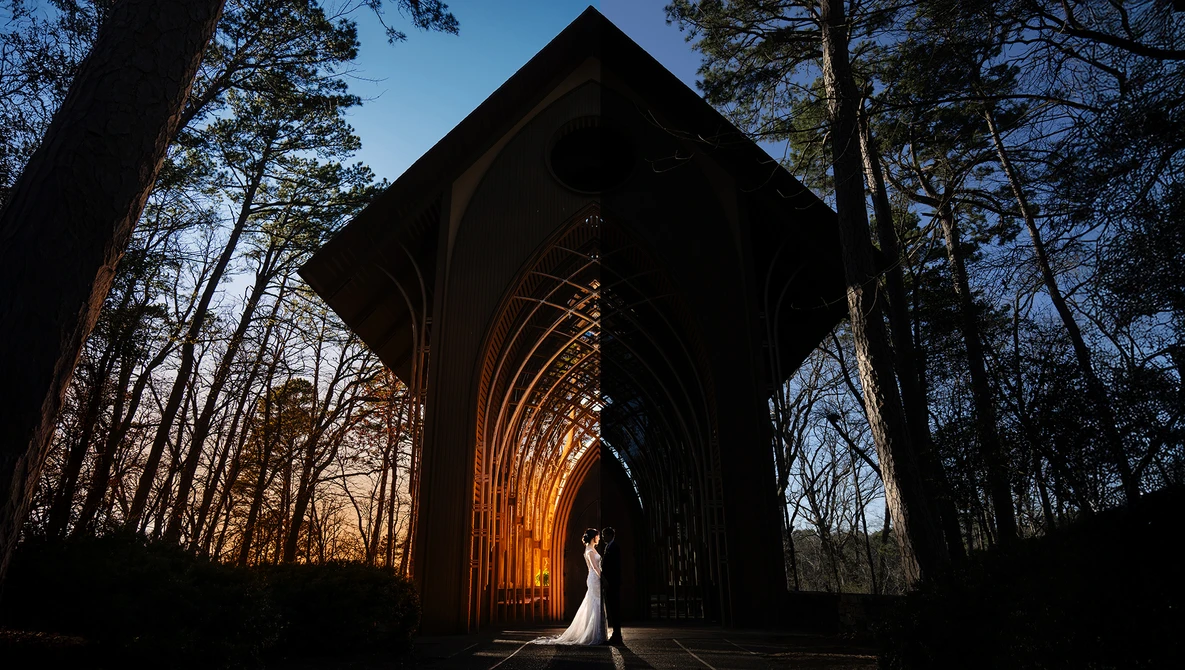 Bride in white dress silhouetted beneath illuminated arched structure surrounded by bare trees at dusk.