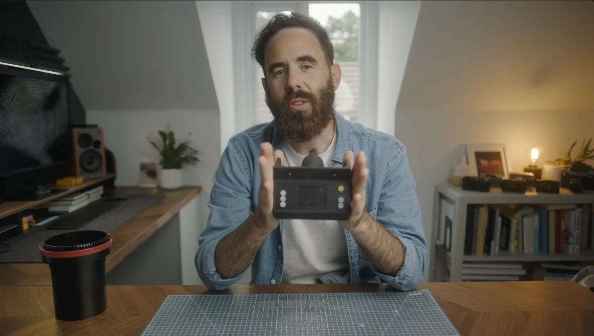 Bearded man holding a camera controller while seated at a desk in a home studio.