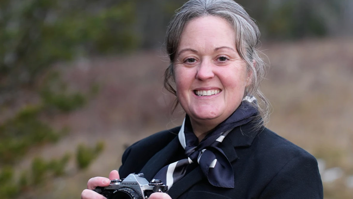 Photographer holding a vintage film camera and smiling at the camera outdoors.
