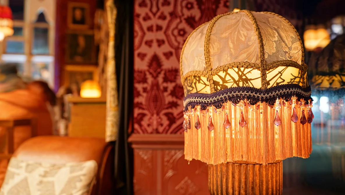 Ornate vintage pendant lamp with cream fabric shade, gold embroidered trim, and long orange beaded fringe hanging in warm interior space.