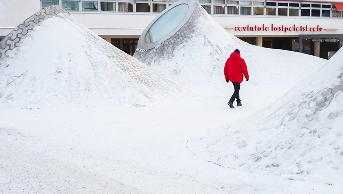 Person in bright red jacket walking across snowy landscape with geometric white structures.