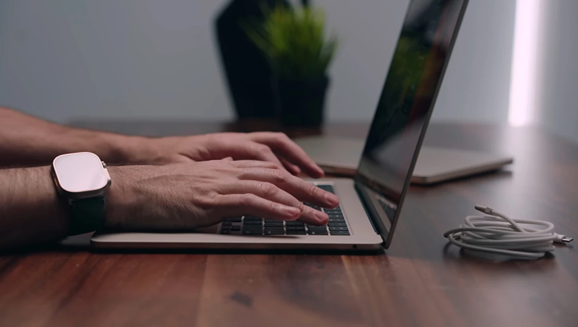 Person typing on laptop keyboard while wearing a smartwatch at a wooden desk.