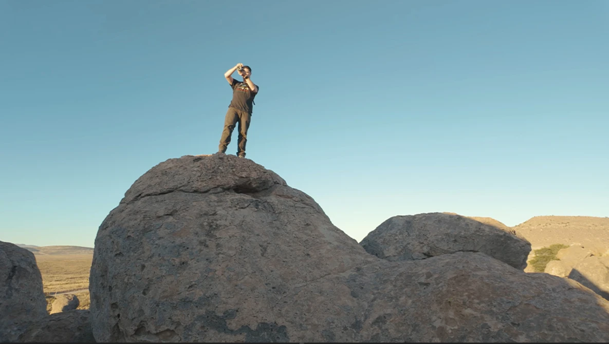 Person standing atop a large rounded boulder against a clear blue sky in an arid landscape.