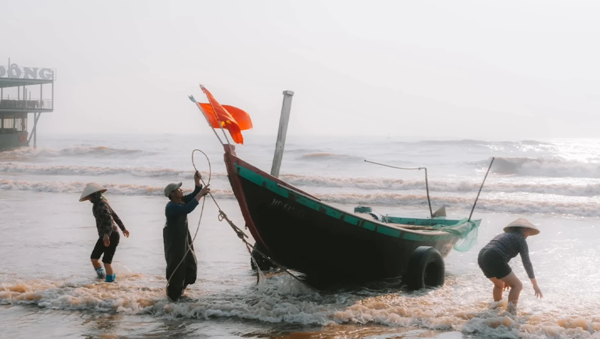 Fishermen in water pulling ropes attached to a green and black wooden boat with an orange flag in rough seas.