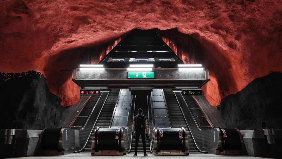 Stockholm subway station with dramatic red rock ceiling and escalators lit by fluorescent panels.