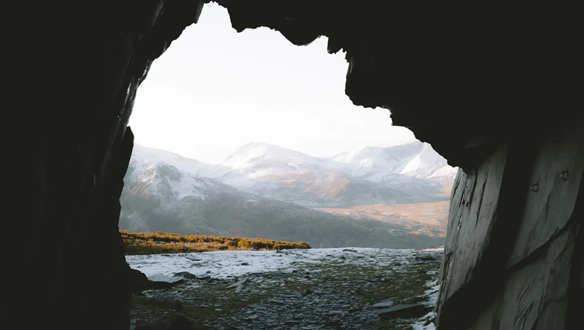 Snow-capped mountains and a rocky coastline viewed through a natural cave opening.