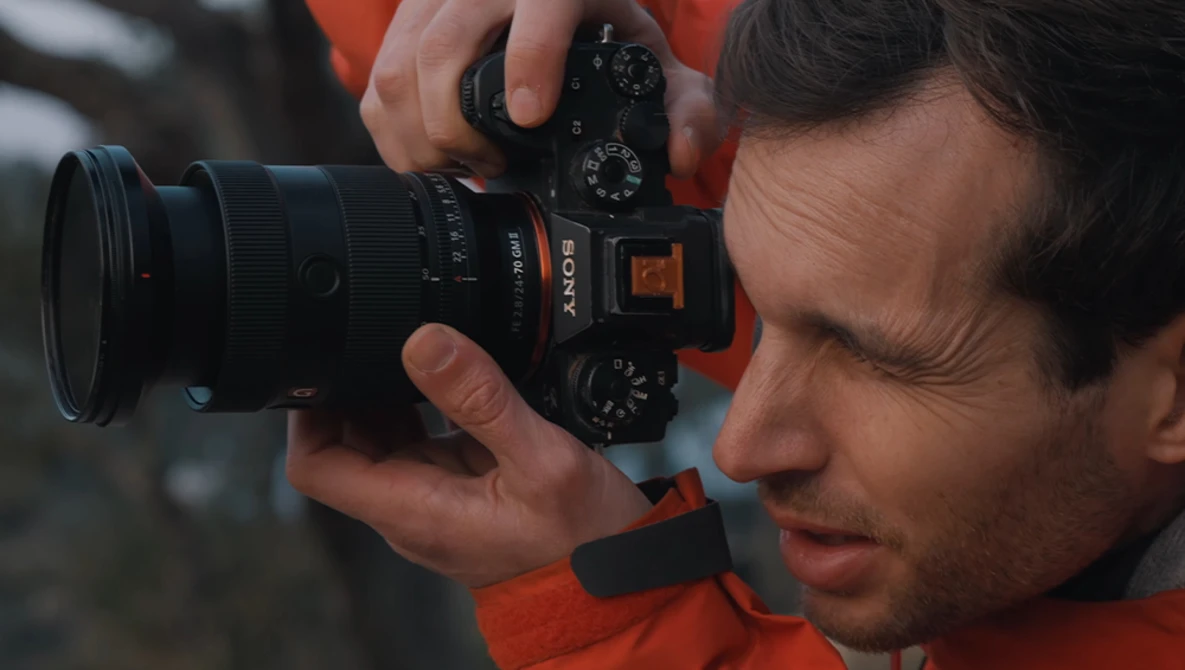 Photographer in orange jacket holding Sony mirrorless camera with telephoto lens to eye.