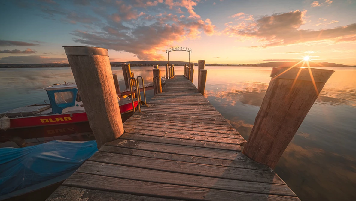 Wooden pier extending into calm water at golden hour sunset, with weathered wooden posts and boats moored alongside.