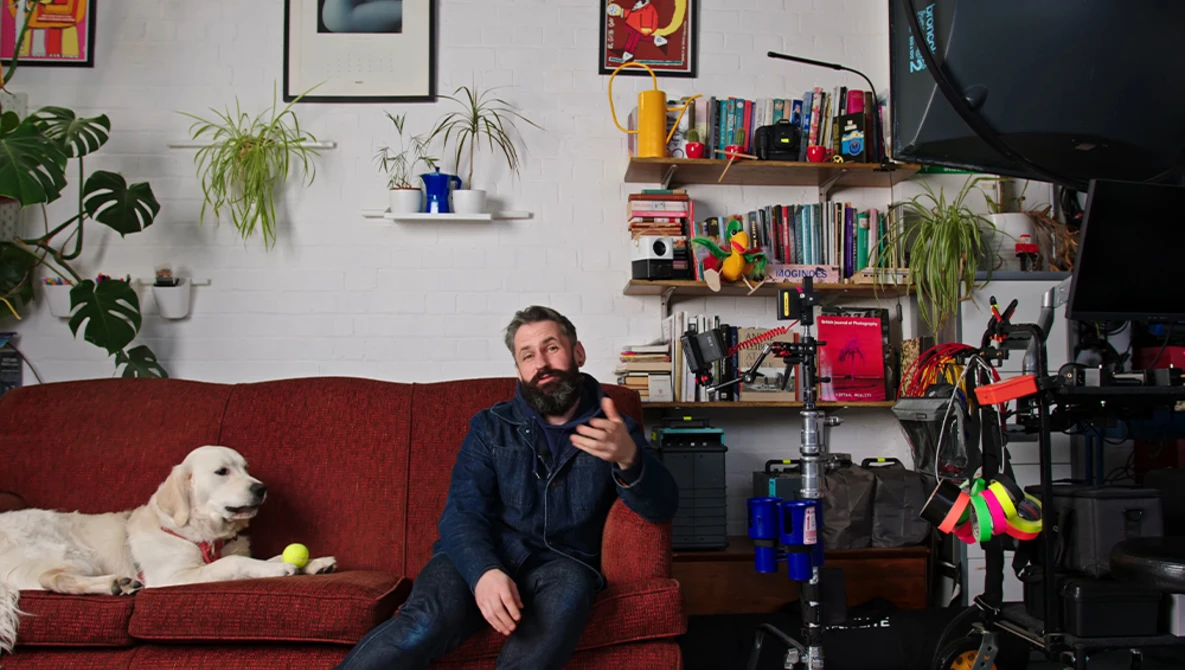 Man sitting on red couch with white dog, gesturing while speaking in a home studio setup.