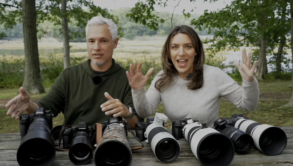 Two people display a collection of camera lenses arranged on a wooden table outdoors.
