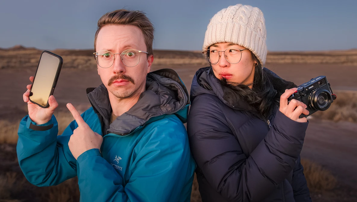 Two photographers in winter clothing posing outdoors with a smartphone and compact camera against a barren landscape.