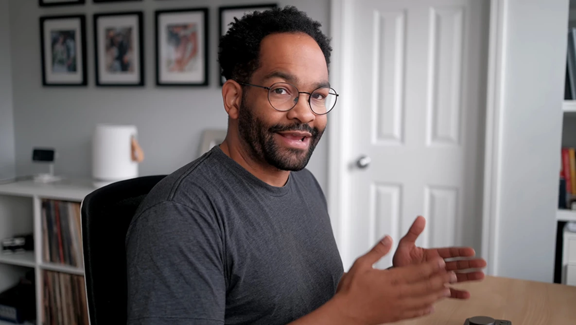 Man wearing glasses and dark t-shirt gesturing while speaking in a home office setting.
