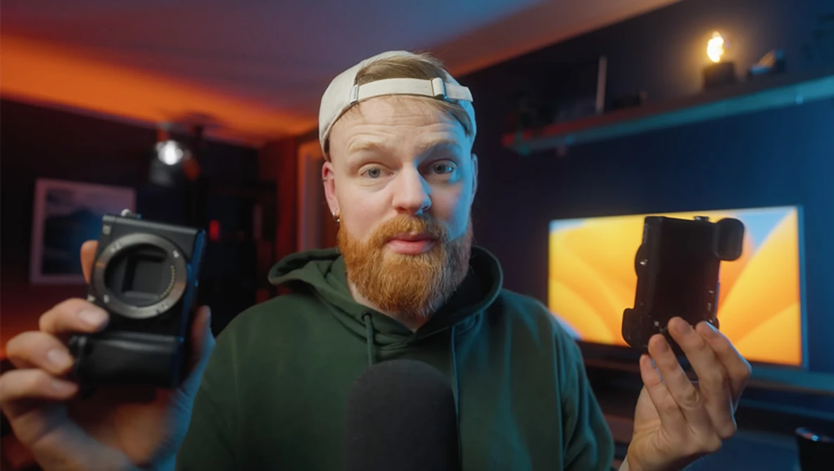 Photographer holding a camera body in one hand and a smartphone in the other, displayed in a studio with warm and cool ambient lighting.