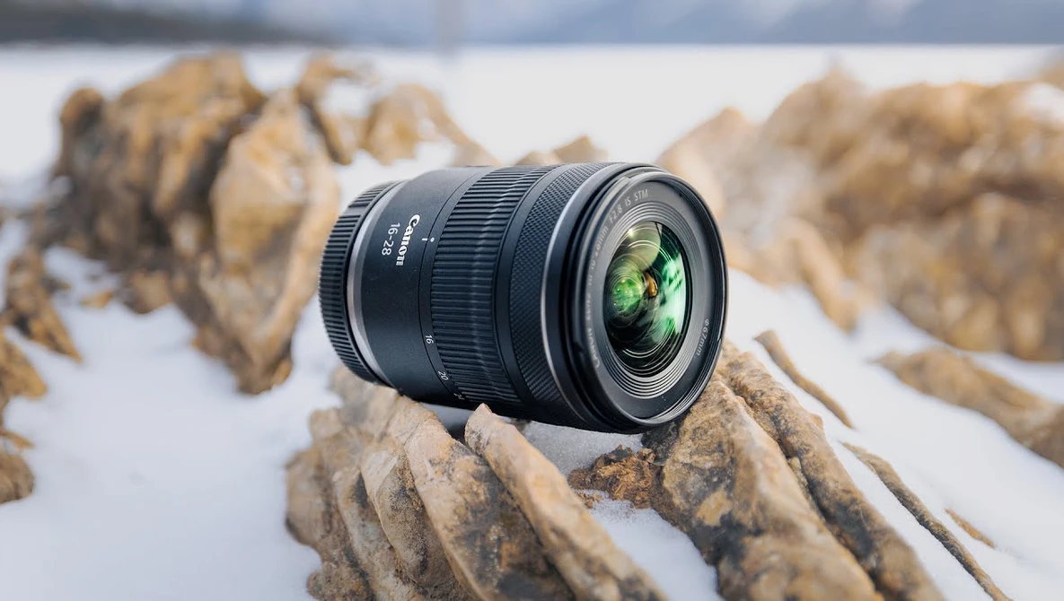 Wide-angle zoom lens positioned on rocky terrain with snowy background and blurred mountains.
