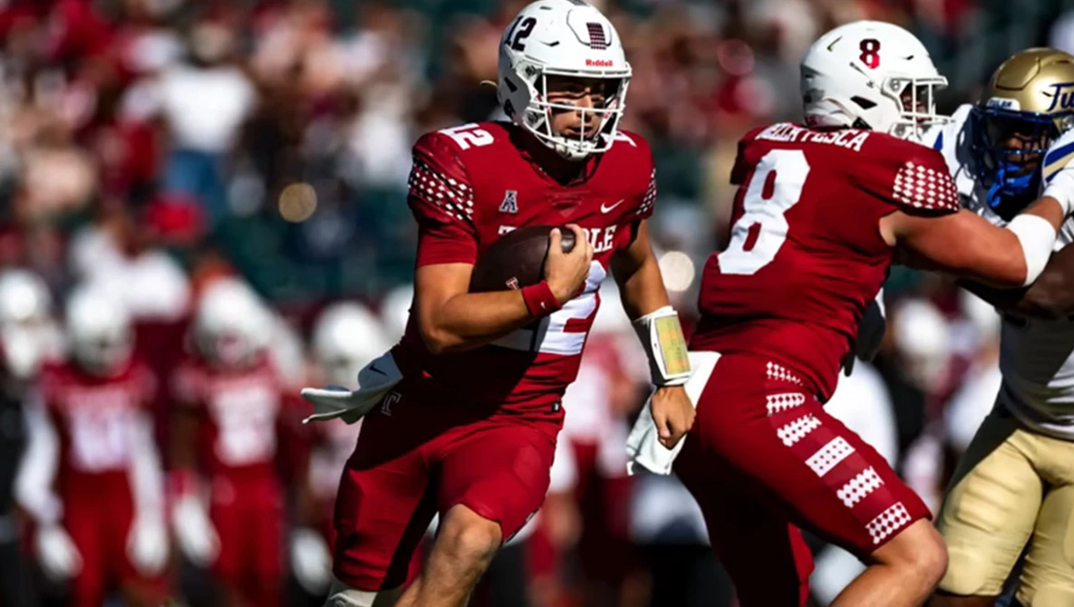 College football quarterback in maroon jersey carries the ball during a game with defensive lineman nearby.