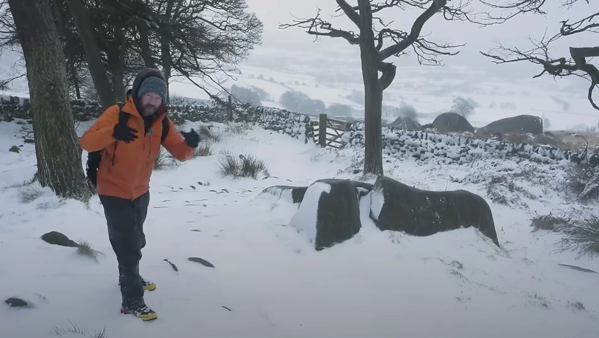 Runner in orange jacket jogging through snowy rural landscape with bare trees and stone walls.