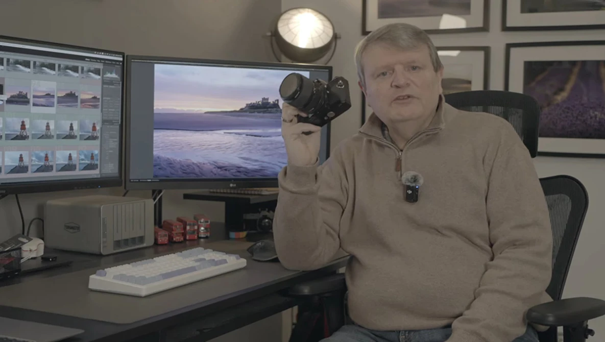 Photographer in studio holding DSLR camera with studio lighting setup visible overhead.