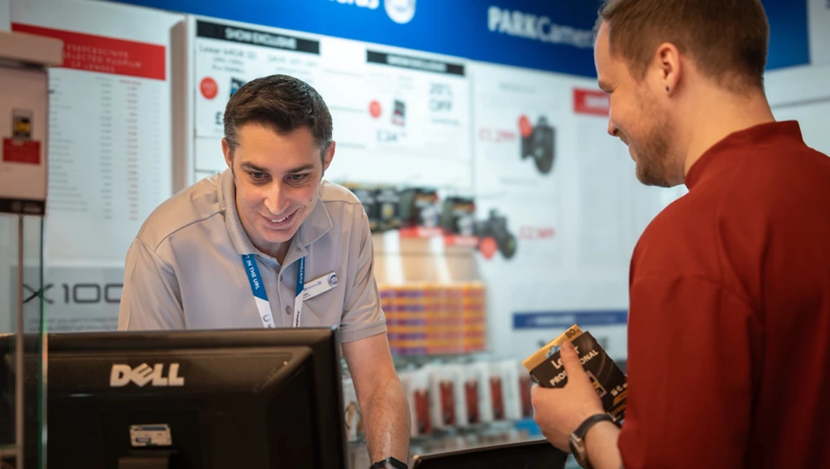 Two men at a tech conference booth discussing equipment, one wearing a lanyard badge at a Dell computer workstation.