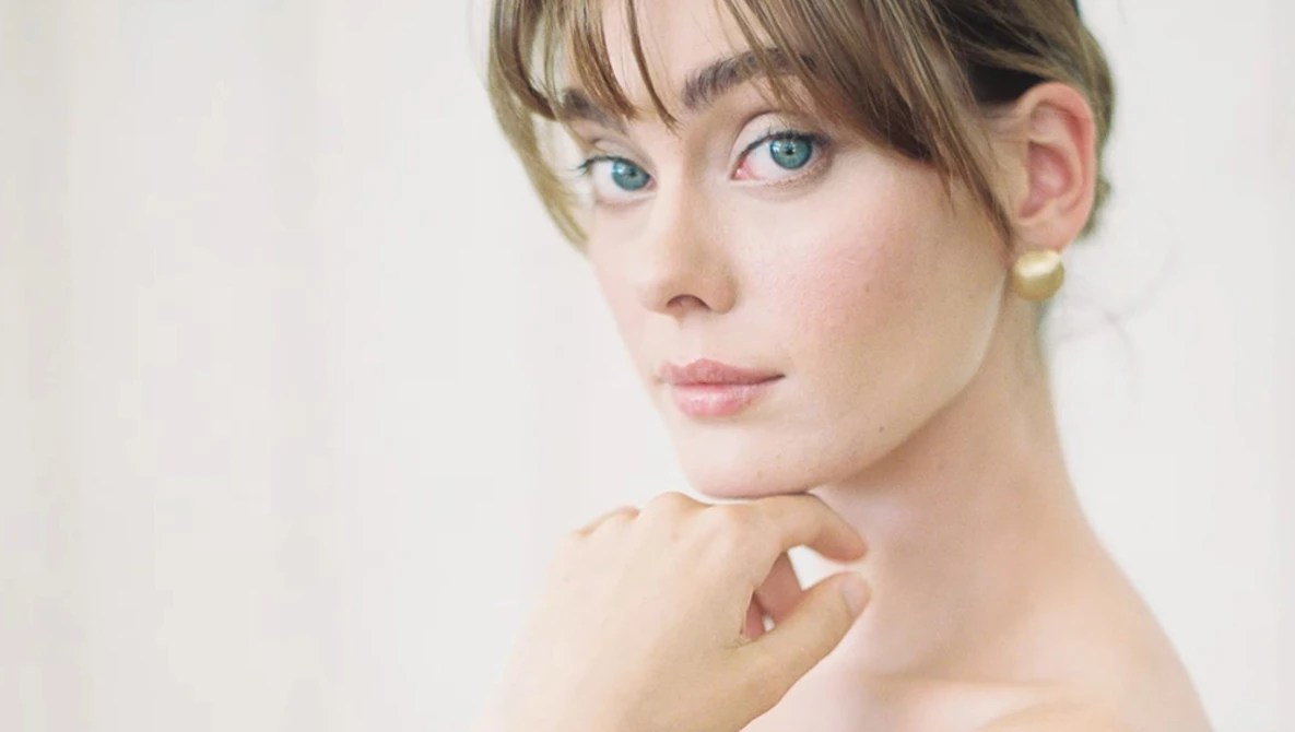 Woman with blue eyes and bangs gazing toward camera with hand near face.