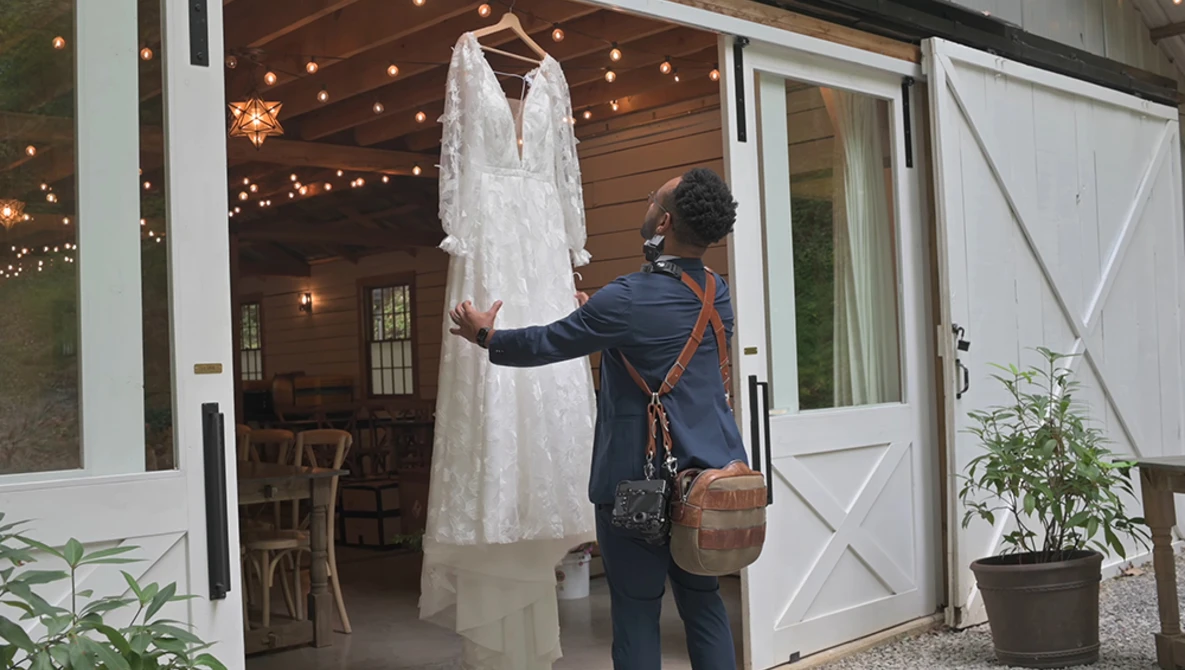 Woman in dark blazer examining a white lace wedding dress hanging indoors.