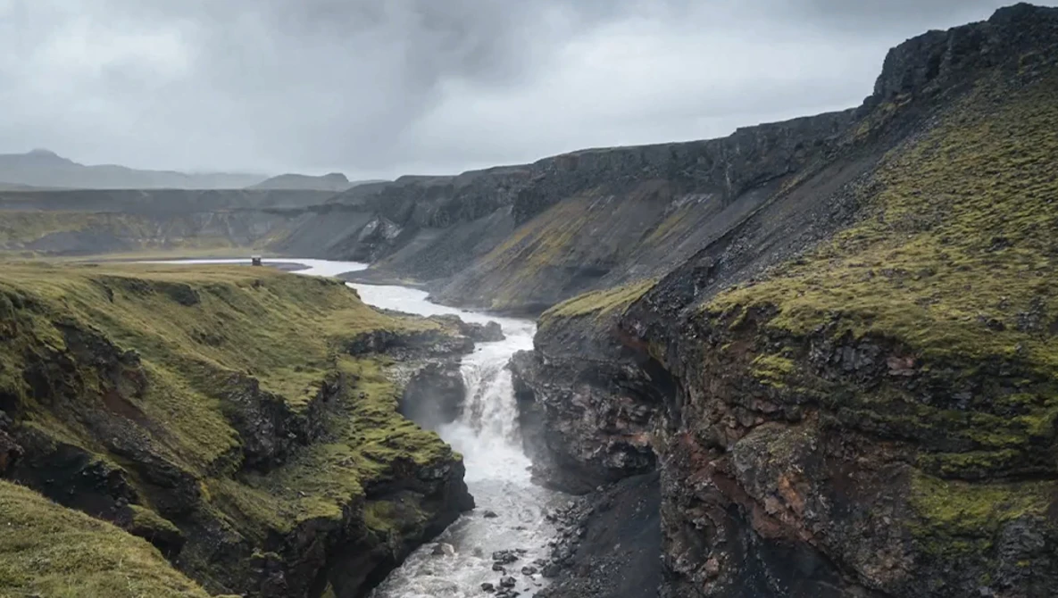 Glacial river cutting through moss-covered canyon with basalt cliffs under overcast sky.