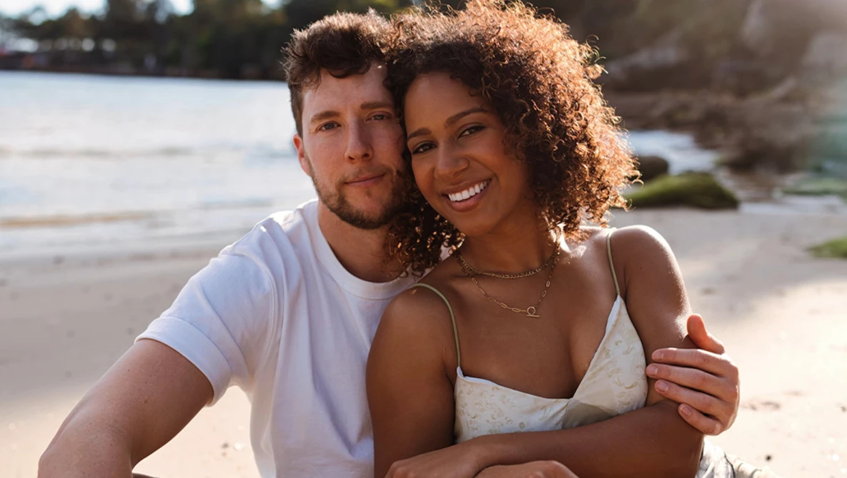 Couple embracing on a sandy beach during golden hour with water and shoreline in background.