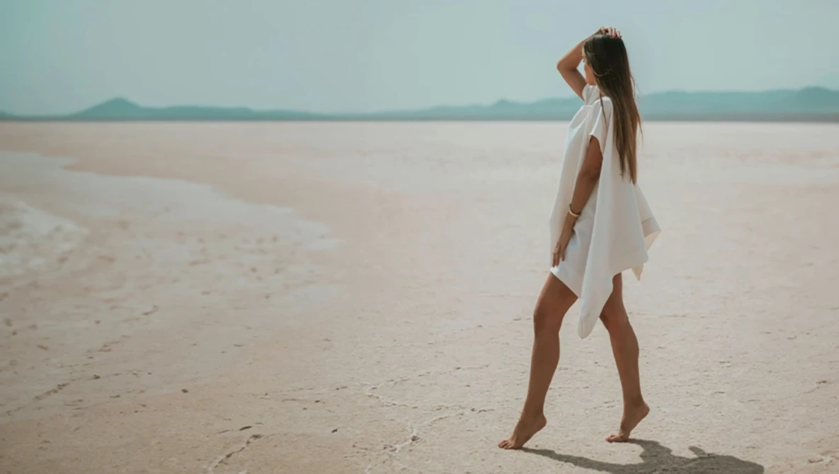Woman in white dress standing barefoot on expansive sandy beach, gazing toward distant horizon and mountains.