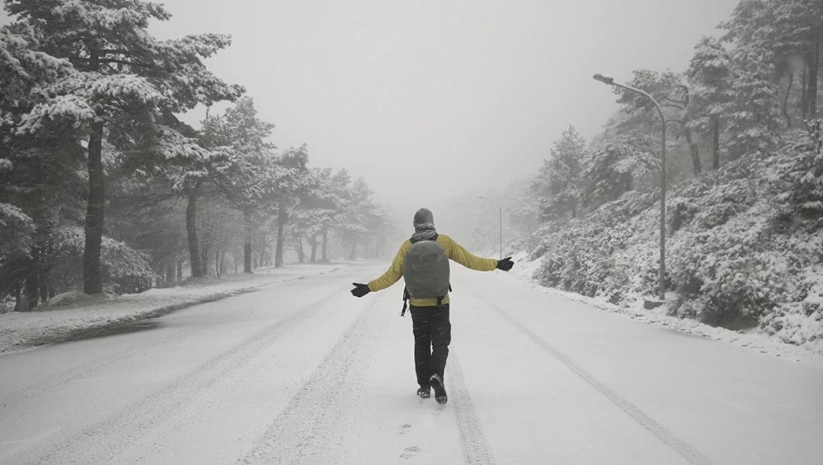 Runner in yellow jacket jogging down a snow-covered tree-lined road with arms outstretched.