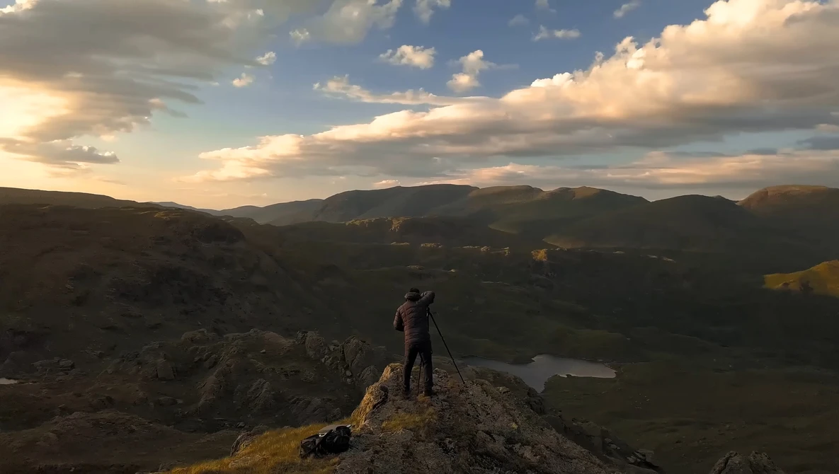 Photographer on rocky outcrop with tripod overlooking mountainous valley at golden hour.