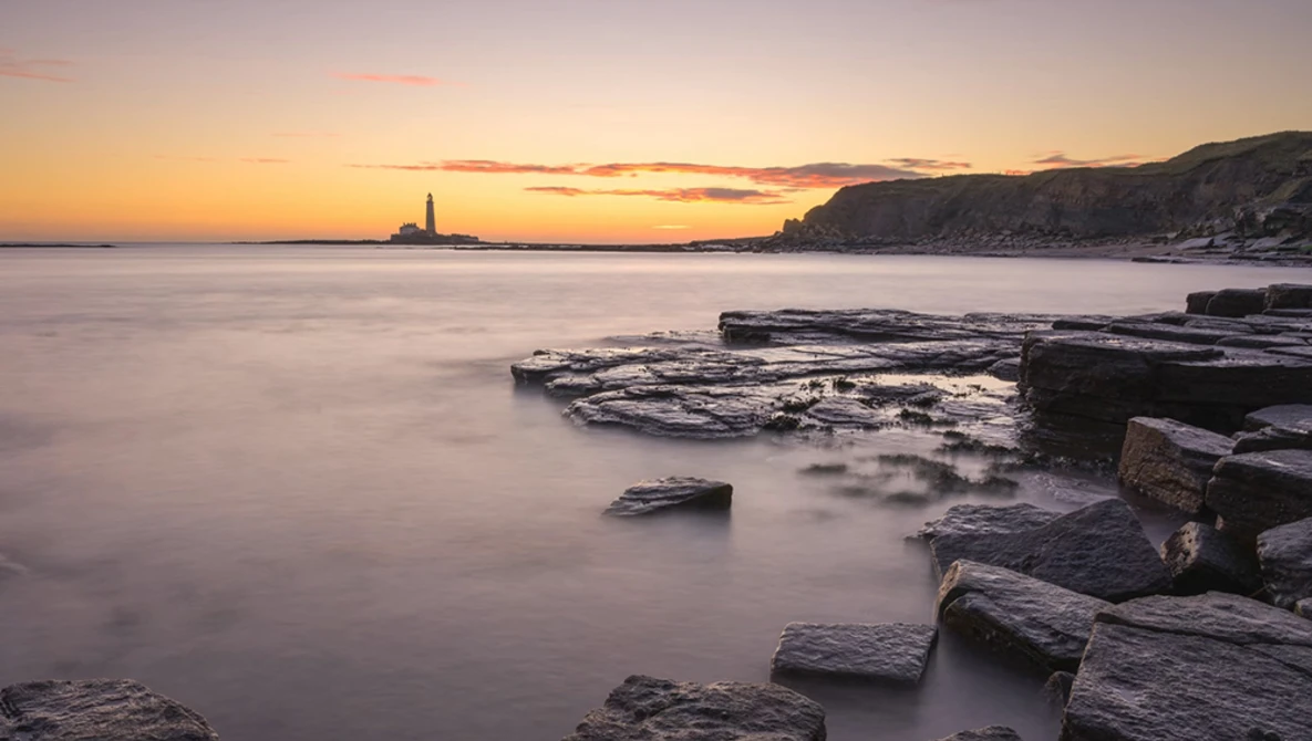 Rocky coastline at sunset with calm water and lighthouse in the distance.