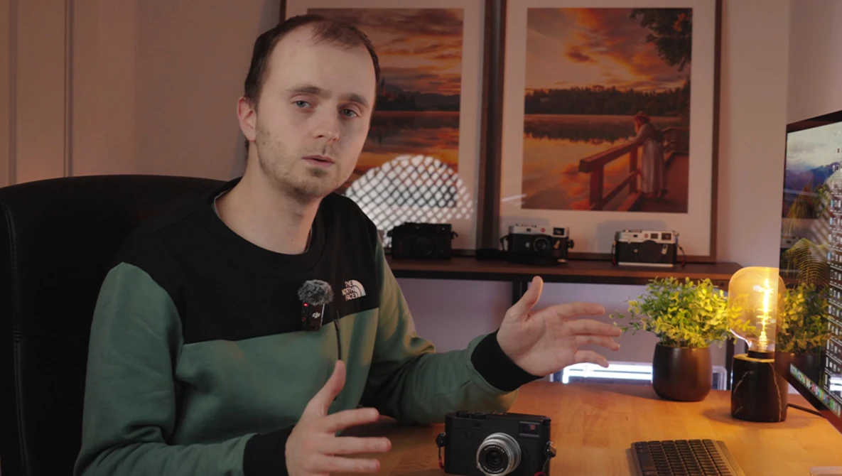 Man in green and black shirt gesturing while speaking at desk with camera and laptop.
