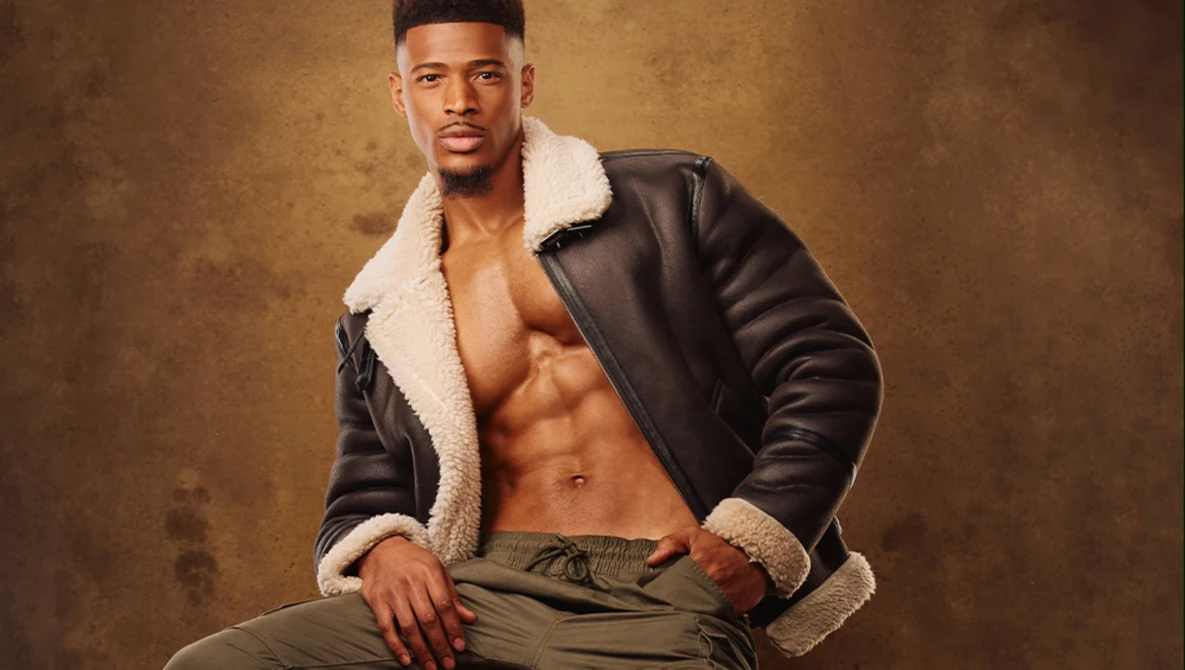 Shirtless man in black leather jacket with sherpa collar posing against textured brown backdrop.