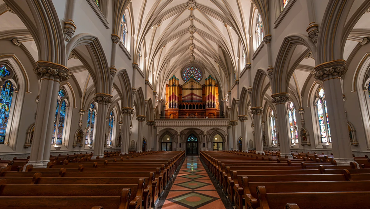 Interior of a Gothic cathedral with soaring vaulted ceilings, ornate pipe organ, and stained glass windows lining both sides.