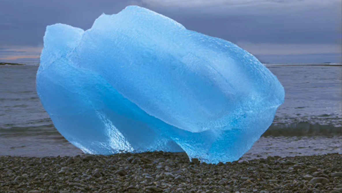 Luminous blue iceberg resting on a rocky beach against a twilight sky.