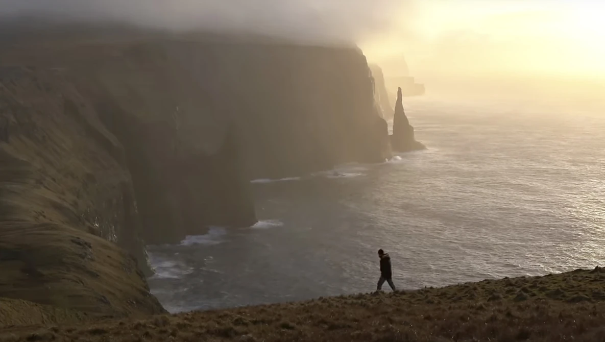 Solitary hiker walking on a cliff edge overlooking dramatic coastal rock formations shrouded in mist at sunrise.