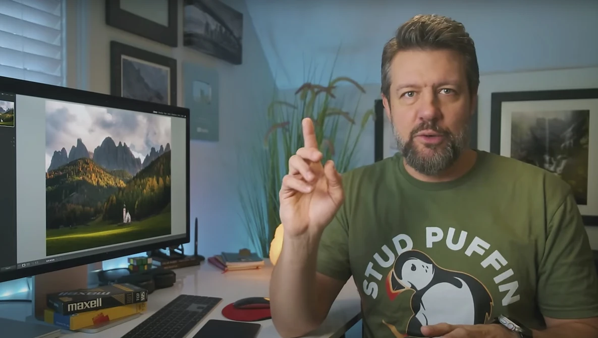 Man in green shirt gesturing while speaking at a desk with a monitor displaying mountain landscape photography.