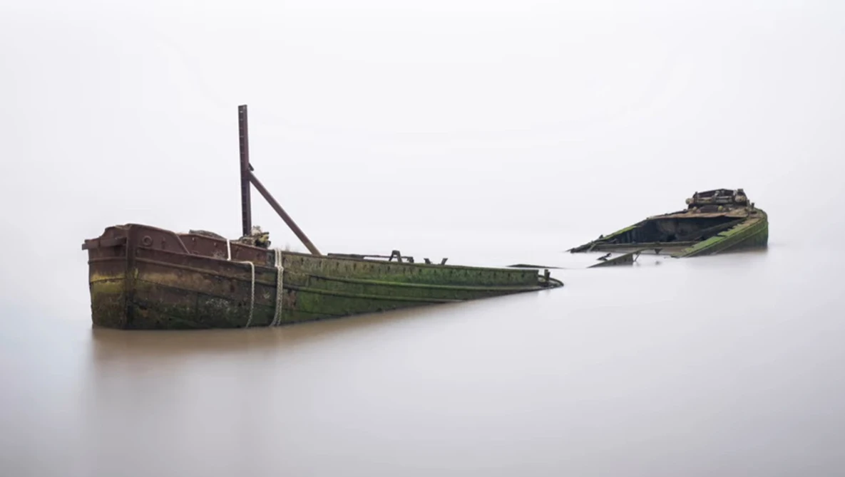 Two rusted, abandoned shipwrecks partially submerged in still water with algae-covered hulls.