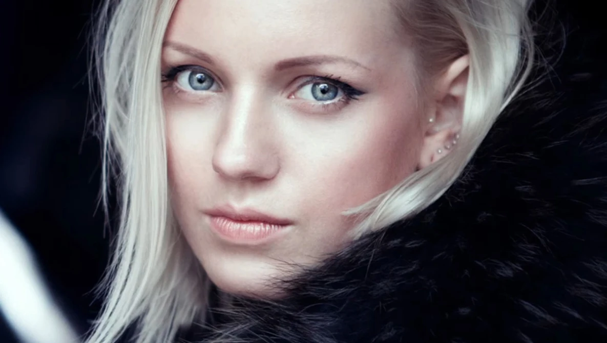 Woman with platinum blonde hair and blue eyes gazing toward camera against dark background.