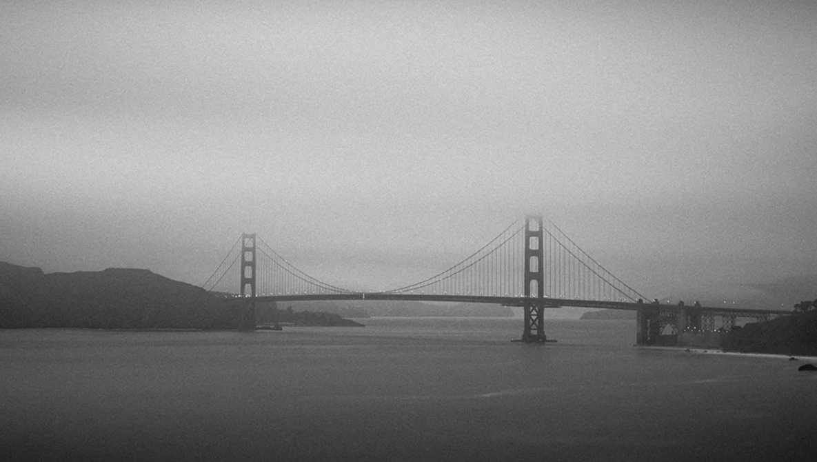 Suspension bridge spanning a body of water under overcast skies in black and white.