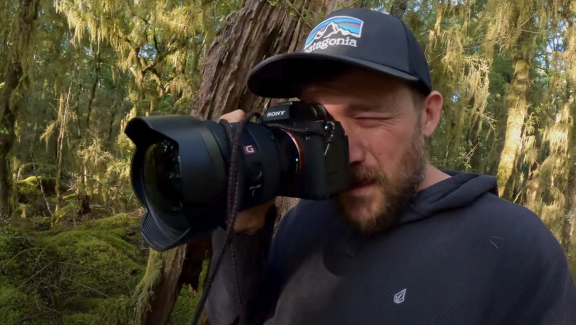 Photographer holding a DSLR camera with a telephoto lens in a mossy forest setting.