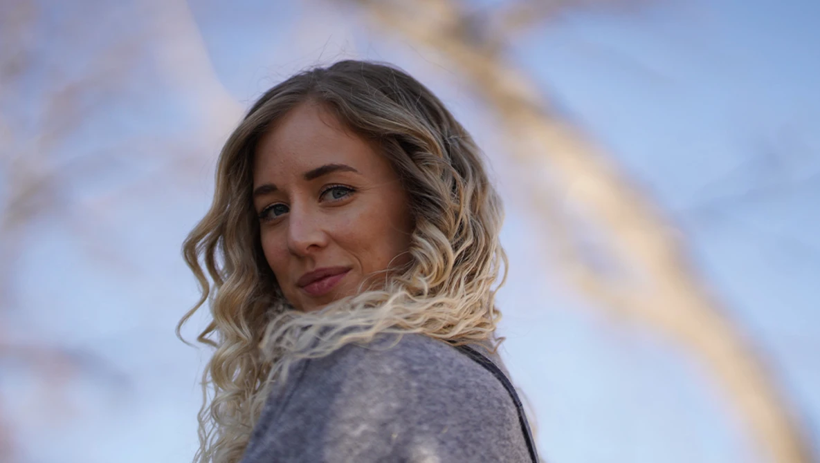 Portrait of a woman with wavy blonde hair wearing a gray sweater, looking over her shoulder against a blurred blue sky background.