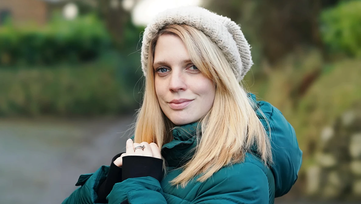 Woman in teal jacket and cream beanie smiling at camera outdoors near water.