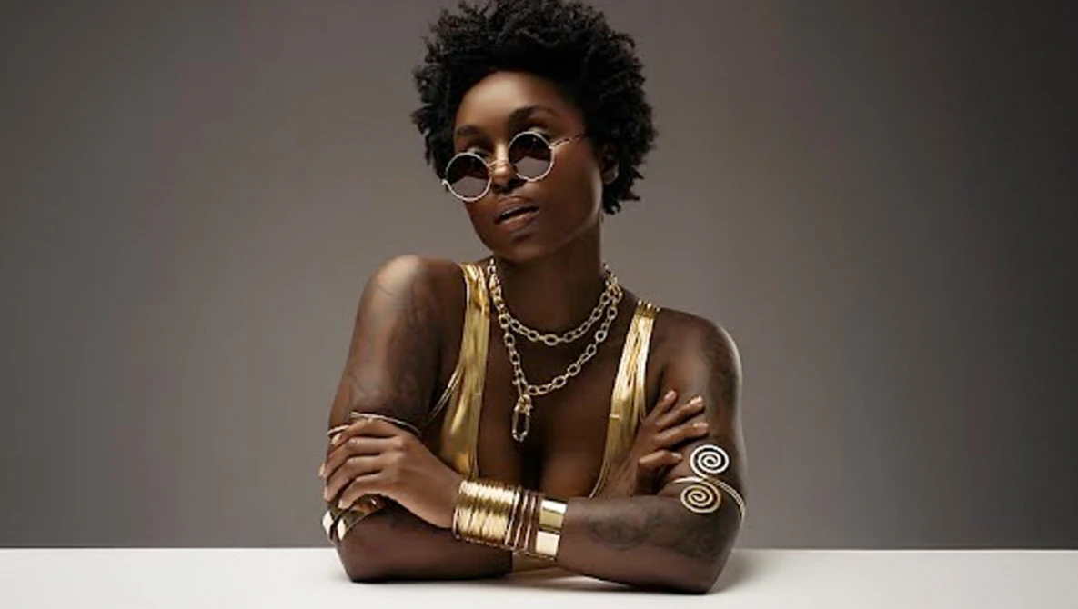 Portrait of a woman wearing round sunglasses, gold jewelry, and metallic body paint against a neutral gray background.