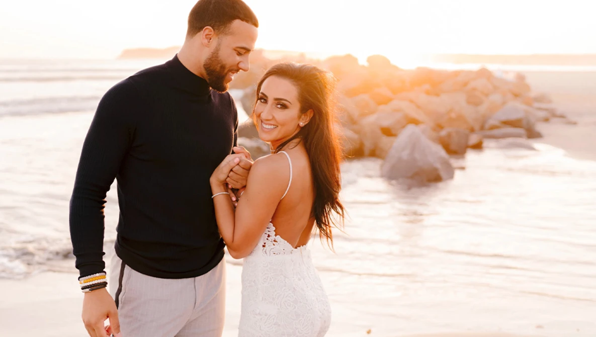 Couple embracing on a beach at sunset with rocky formations in the background.