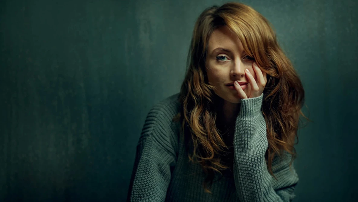 Woman with long brown hair resting her face against her hand, wearing a gray sweater against a teal background.