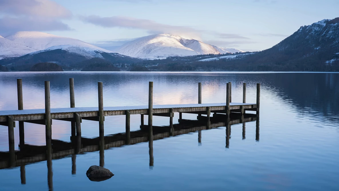 Wooden dock extending into still lake with snow-capped mountains reflected in water at dawn.