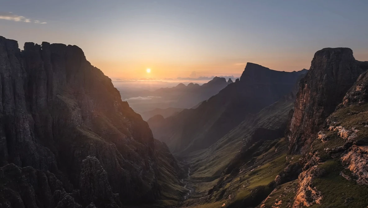 Sunrise over a mountain valley with dramatic cliff faces framing a distant sun breaking through layers of mist.